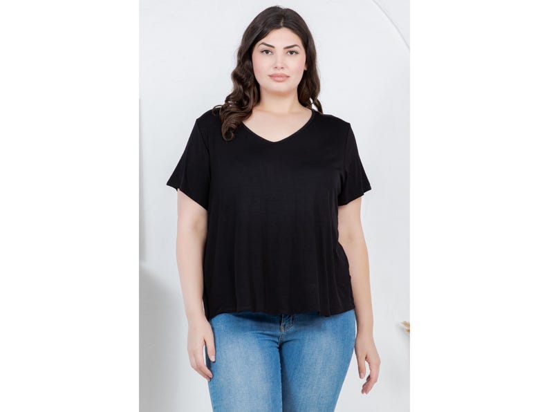 Op de afbeelding: Een vrouw draagt een zwart V-hals T-shirt en een blauwe spijkerbroek. De top met korte mouwen heeft een losse pasvorm. De jeans is licht gewassen denim. De achtergrond is een effen witte muur.