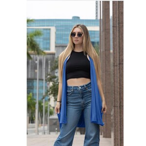Pode incluir: Uma mulher vestindo um cardigã azul sobre um top preto e jeans azul. Ela está usando óculos de sol redondos e tem cabelo loiro comprido.