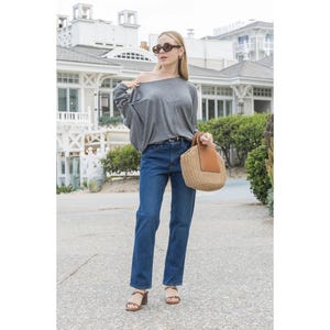 May include: A woman wearing a gray off-the-shoulder top, blue jeans, and brown sandals. She carries a woven straw handbag with a brown leather handle. The background features white buildings and greenery.