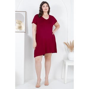 Puede incluir: Vestido burdeos con cuello en V y mangas cortas. El vestido tiene un dobladillo ligeramente asim&eacute;trico y llega a la rodilla. La modelo lleva sandalias transparentes. El fondo es blanco con una obra de arte enmarcada y una peque&ntilde;a mesa blanca.