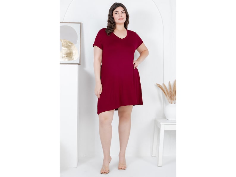 Puede incluir: Vestido burdeos con cuello en V y mangas cortas. El vestido tiene un dobladillo ligeramente asim&eacute;trico y llega a la rodilla. La modelo lleva sandalias transparentes. El fondo es blanco con una obra de arte enmarcada y una peque&ntilde;a mesa blanca.