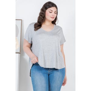Op de afbeelding: Een vrouw draagt een lichtgrijs V-hals T-shirt en een blauwe jeans. Het T-shirt heeft korte mouwen en een iets langere zoom aan de achterkant. De jeans heeft een klassieke blauwe wassing.