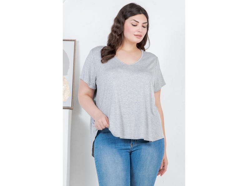 Op de afbeelding: Een vrouw draagt een lichtgrijs V-hals T-shirt en een blauwe jeans. Het T-shirt heeft korte mouwen en een iets langere zoom aan de achterkant. De jeans heeft een klassieke blauwe wassing.