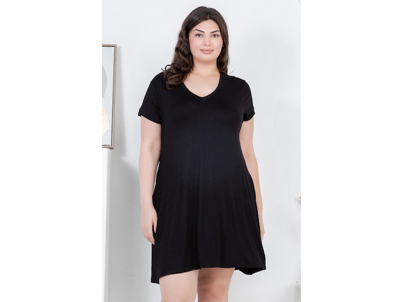 Puede incluir: Un vestido negro con cuello en V y mangas cortas. El vestido tiene un dise&ntilde;o sencillo y fluido y llega hasta la rodilla. La modelo lleva el vestido en un estudio con un fondo neutro.