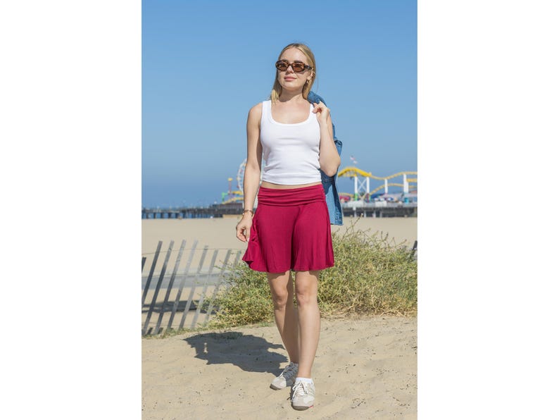 Op de afbeelding: Een vrouw draagt een witte tanktop en een korte, uitlopende, rode rok. Ze draagt ook een zonnebril en witte sneakers. Een spijkerjasje ligt over haar schouder. De achtergrond toont een strand, de zee en een pier met een reuzenrad.