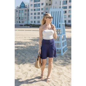 Op de afbeelding: Een vrouw draagt een witte strapless top en een marineblauwe rok, met een geweven handtas. Ze staat op een zandstrand met een strandwachtpost op de achtergrond. De gebouwen zijn wit en blauw.