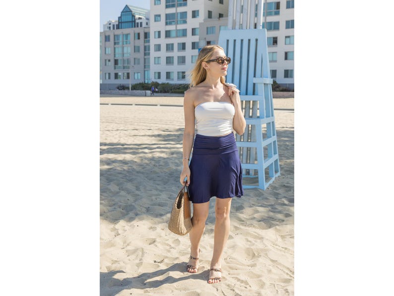 Op de afbeelding: Een vrouw draagt een witte strapless top en een marineblauwe rok, met een geweven handtas. Ze staat op een zandstrand met een strandwachtpost op de achtergrond. De gebouwen zijn wit en blauw.