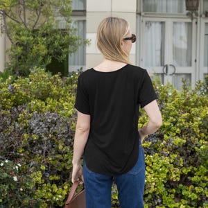 Op de afbeelding: Een zwart T-shirt met korte mouwen en een ronde hals met een gebogen zoom. De persoon draagt een blauwe spijkerbroek en een zonnebril. Een bruine leren tas wordt in de hand gehouden. De achtergrond toont groene struiken en een gebouw.