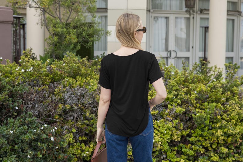 Op de afbeelding: Een zwart T-shirt met korte mouwen en een ronde hals met een gebogen zoom. De persoon draagt een blauwe spijkerbroek en een zonnebril. Een bruine leren tas wordt in de hand gehouden. De achtergrond toont groene struiken en een gebouw.