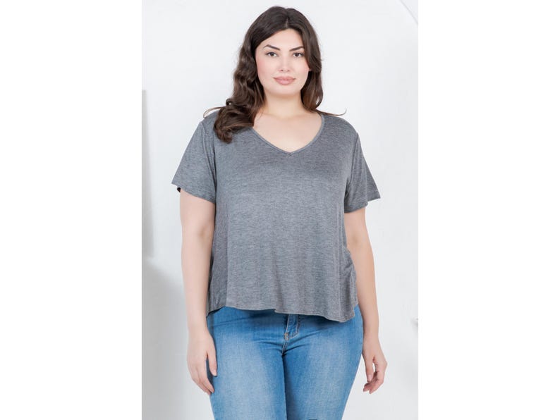 Op de afbeelding: Een grijs V-hals T-shirt met korte mouwen. Het shirt wordt gecombineerd met lichtblauwe jeans. Het model staat voor een witte achtergrond. Het shirt heeft een relaxte pasvorm.