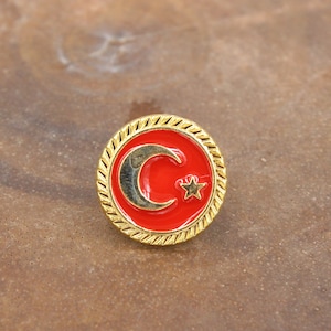 Könnte beinhalten: Eine goldfarbene Anstecknadel mit einem roten, emaillierten Halbmond und einem weißen Stern in der Mitte.