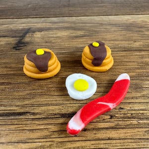 May include: A miniature breakfast scene featuring two stacks of pancakes with chocolate syrup and a pat of butter, a fried egg, and a red and white curved piece of bacon. The items are arranged on a wooden surface.