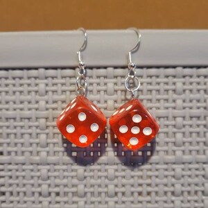 May include: A pair of red dice dangle from silver earring hooks. Each die has white dots on each side.