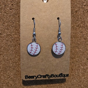 May include: A pair of silver dangle earrings with a white baseball design. The baseball has red stitching and is on a silver circular base.
