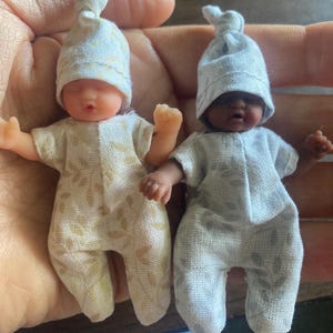 May include: Two miniature baby dolls with vinyl skin, each wearing a matching fabric onesie and a knotted cap. One doll has light skin with a cream and gold patterned outfit, while the other has dark skin with a light blue and grey patterned outfit.