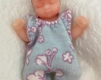 May include: A small, sleeping baby doll wearing a light blue and purple floral onesie with a white headband. The doll has closed eyes and a realistic appearance.