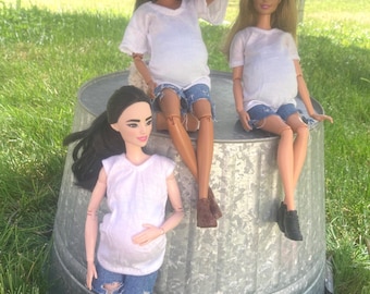 May include: Three fashion dolls, each wearing a white top and denim shorts, are posed on a galvanized metal container. The dolls have different skin tones and hairstyles. The dolls appear to be pregnant.