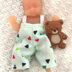 May include: A small doll wearing light green pants with colorful triangle patterns, and white suspenders. A brown teddy bear figurine with a small hat is next to the doll. The doll is lying on a white, textured surface.
