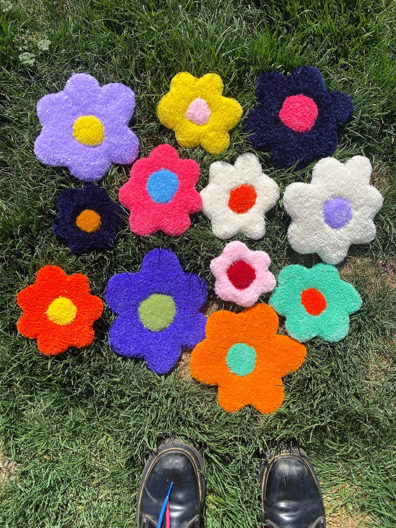 May include: A collection of 12 colorful, fluffy, flower-shaped rugs with different colored centers. The rugs are arranged in a heart shape on a green grassy lawn.