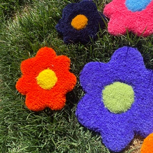 May include: Five colorful flower-shaped plush cushions in various colors, including orange, blue, pink, purple, and black, with yellow, green, and blue centers. The cushions are arranged on a bed of green grass.
