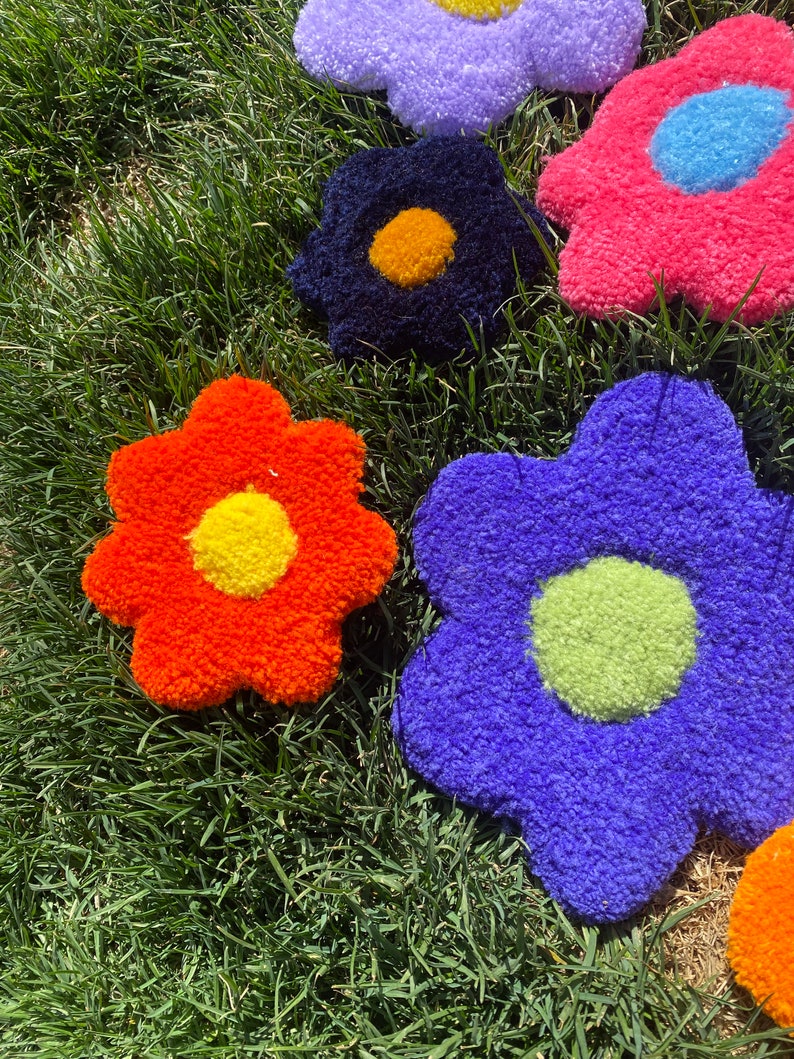 May include: Five colorful flower-shaped plush cushions in various colors, including orange, blue, pink, purple, and black, with yellow, green, and blue centers. The cushions are arranged on a bed of green grass.