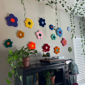 May include: A white wall decorated with colorful felt flower wall decor. The flowers are in various shades of pink, blue, yellow, orange, and green. There are also some green vines hanging from the ceiling.