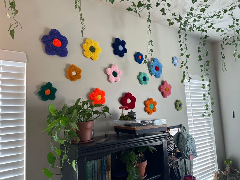 May include: A white wall decorated with colorful felt flower wall decor. The flowers are in various shades of pink, blue, yellow, orange, and green. There are also some green vines hanging from the ceiling.
