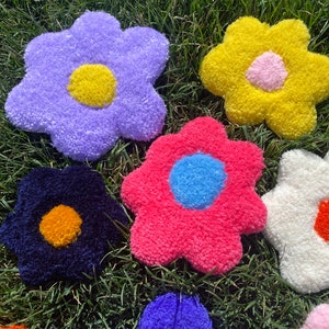 May include: Six colorful plush flower-shaped cushions in various colors, including purple, yellow, pink, blue, white, and black, with contrasting colored centers. The cushions are arranged on a bed of green grass.