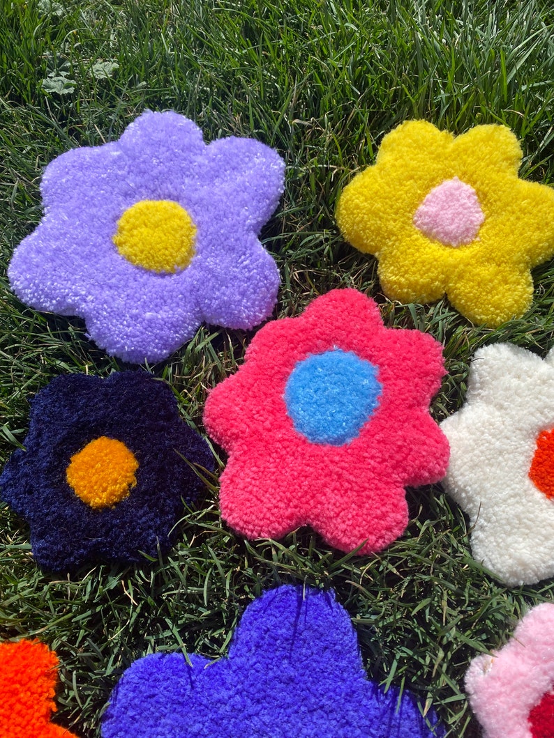 May include: Six colorful plush flower-shaped cushions in various colors, including purple, yellow, pink, blue, white, and black, with contrasting colored centers. The cushions are arranged on a bed of green grass.