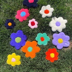 May include: Ten colorful, fluffy, flower-shaped rugs in various colors, including pink, blue, green, orange, yellow, and white, are arranged on a green grassy lawn.