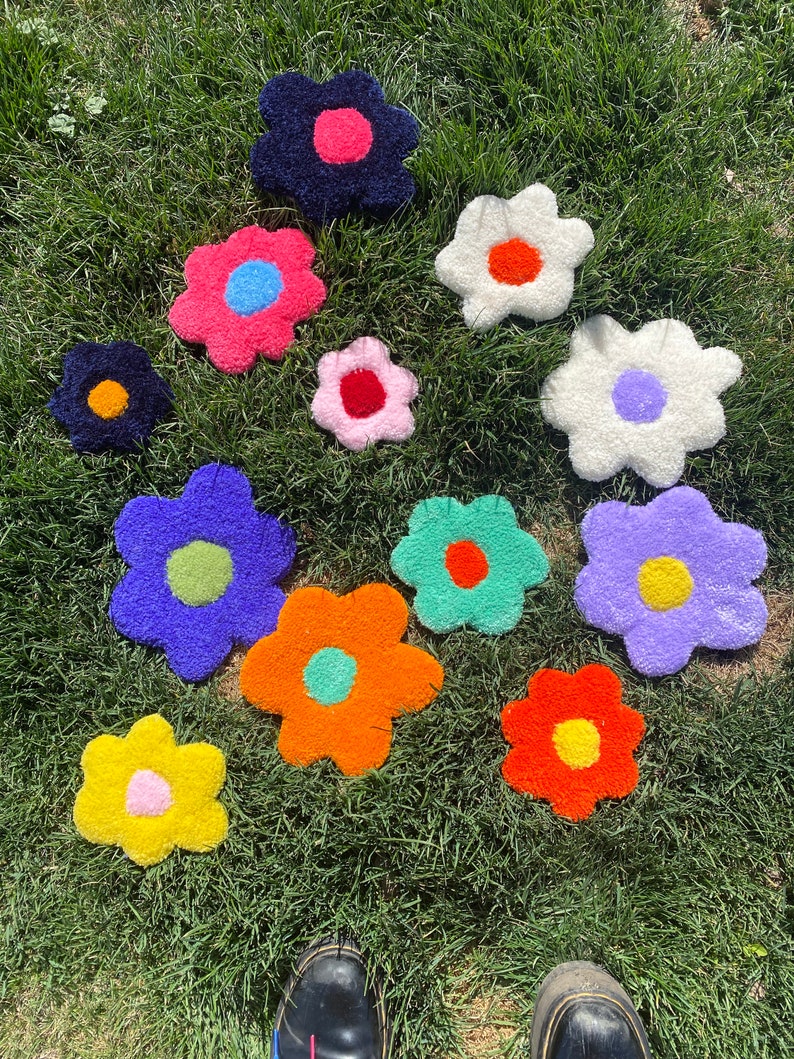 May include: Ten colorful, fluffy, flower-shaped rugs in various colors, including pink, blue, green, orange, yellow, and white, are arranged on a green grassy lawn.
