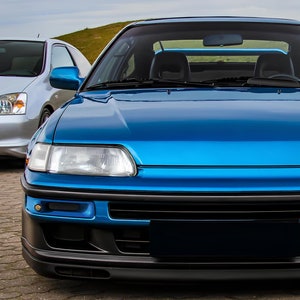 Peut inclure: Une Honda CRX bleue avec un pare-chocs avant noir et une calandre noire. La voiture est garée sur une surface pavée.