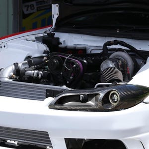 May include: A white sports car with the hood open, revealing a high-performance engine bay with a turbocharger, a purple cam gear, and metallic piping. The front features carbon fiber headlight housings and a large front-mounted intercooler.