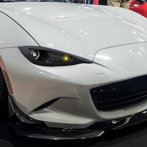 Puede incluir: Un coche deportivo de color gris claro con un parachoques delantero bajo con un divisor negro, faros estrechos y angulares con una pequeña luz amarilla y una gran parrilla negra. El vehículo tiene ruedas anchas de radios metálicos.