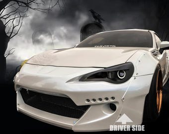 May include: A white sports car with a wide body kit, black headlight housings, and a front bumper featuring a black mesh grille and five small circular lights. The windscreen displays the text TAILORED and the lower bumper says DRIVER SIDE.