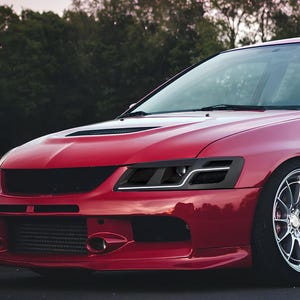 Puede incluir: Un Mitsubishi Lancer Evolution rojo con faros delanteros negros modificados y una parrilla negra. El coche tiene una llanta de aleación plateada y un parachoques delantero negro.