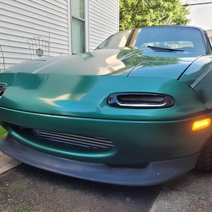 Può includere: Una Mazda MX-5 verde con fari neri e un paraurti anteriore nero. L'auto ha una griglia nera e uno spoiler nero.