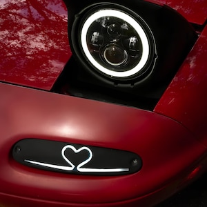May include: A red car with a black headlight and a white ring around the lens. The car has a black grill with a white heart-shaped design.