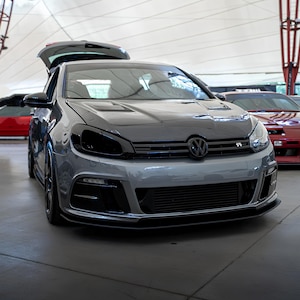 Puede incluir: Un Volkswagen Golf plateado con un capó negro y faros oscurecidos. El coche tiene un parachoques delantero personalizado con una parrilla negra y un alerón delantero negro.
