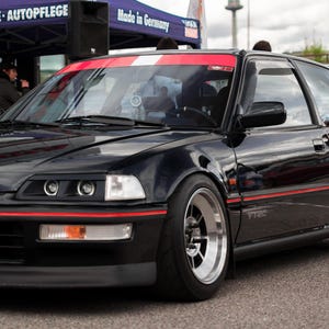 May include: A black Honda Civic with a red and white stripe across the hood. The car has a black bumper and black wheels with silver rims. The car has a license plate that reads "179".