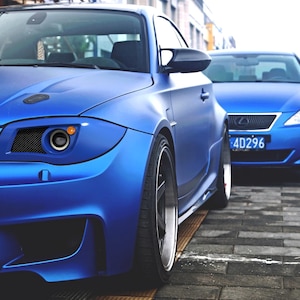 Puede incluir: Dos coches azules estacionados sobre una superficie de ladrillo. El coche en primer plano tiene un acabado mate, ruedas negras y un diseño de faros distintivo. El segundo coche también es azul, con una matrícula visible.