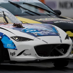 Może przedstawiać: Biały samochód sportowy z niebiesko-żółtym wzorem. Samochód ma duży przedni grill i spojler. Samochód znajduje się na torze i zaraz skręci.