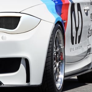Puede incluir: Un BMW Serie 1 blanco con una franja de carreras en blanco y negro y el texto "CYC Group" y "Sabelt" en el lateral. El coche tiene un divisor delantero negro y un difusor trasero negro.