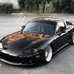 May include: A black Mazda MX-5 with a hardtop roof, aftermarket wheels, and a lowered suspension. The car is parked in an empty car park.