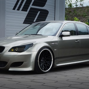 Może przedstawiać: Srebrno-szary sedan BMW serii 5 z przyciemnionymi reflektorami i felgami posprzedażnymi. Samochód ma niestandardowy zestaw nadwozia z dolnym zderzakiem przednim i progami bocznymi.