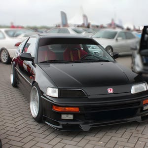 May include: A black Honda CRX sports car with a red interior and silver rims is parked on a brick surface. The car has a front spoiler and a Honda emblem. Other cars and people are in the background.