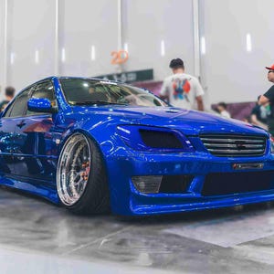 May include: A bright blue car with blacked-out headlights and a black grille. The car is parked in a large, empty space with people standing around it.