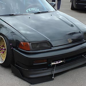 May include: A black Honda Civic with a custom body kit, gold wheels, and a black mesh grille. The car has a large front spoiler and a small rear spoiler.