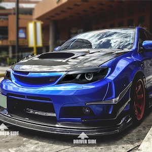 May include: A blue sedan featuring a carbon fiber hood with a central scoop, dark-tinted headlights, and a carbon fiber front splitter. The vehicle includes red wheels and white text labels pointing to the passenger side and driver side.