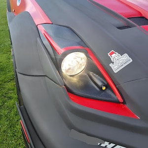Puede incluir: Primer plano de un coche personalizado rojo y negro con un capó de fibra de carbono y una pegatina "1458 Custom Parts". El coche tiene un faro redondo grande y una pegatina de neumático "Michelin" en blanco y negro.